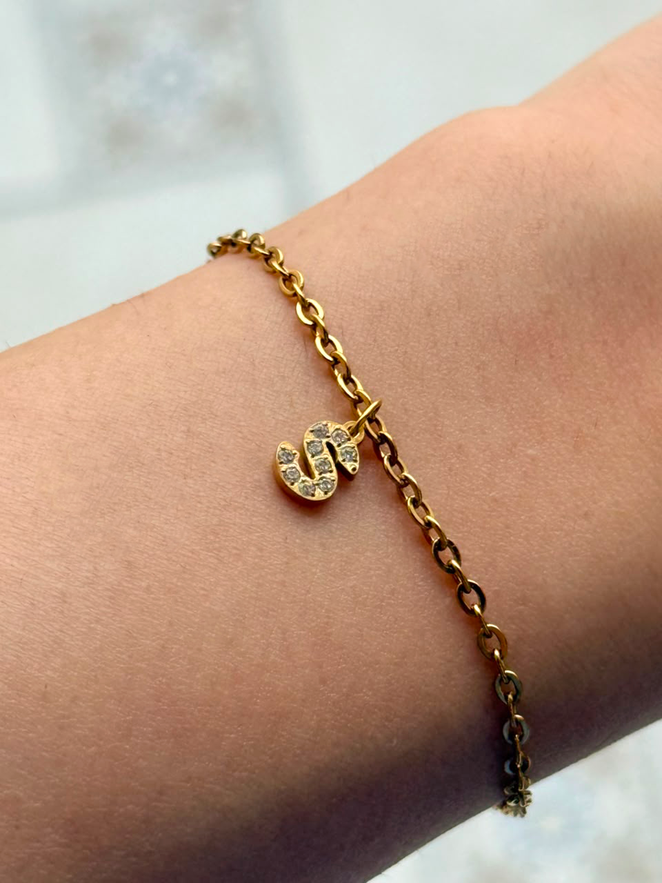 A delicate gold chain bracelet with a small initial pendant paved with clear stones, shown worn on a person's wrist.