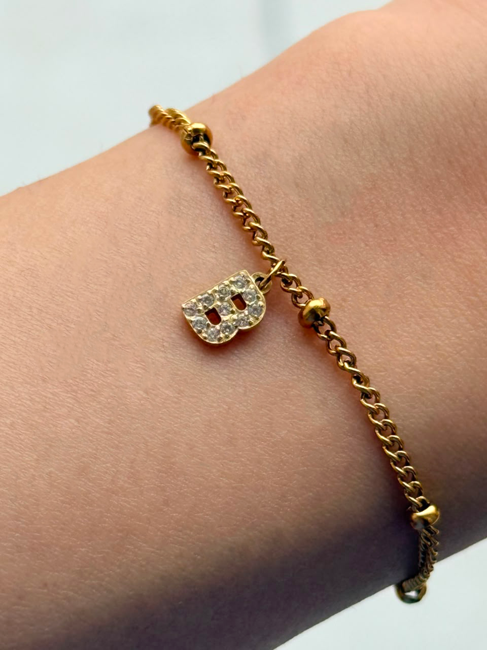 A gold chain bracelet with a small initial 'B' charm encrusted with clear stones, shown worn on a person's wrist.