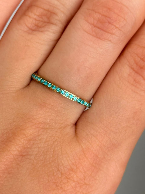 A close-up of a delicate gold eternity ring band set with small round-cut turquoise gemstones worn on a finger. 14K gold-plated, 316L stainless steel
