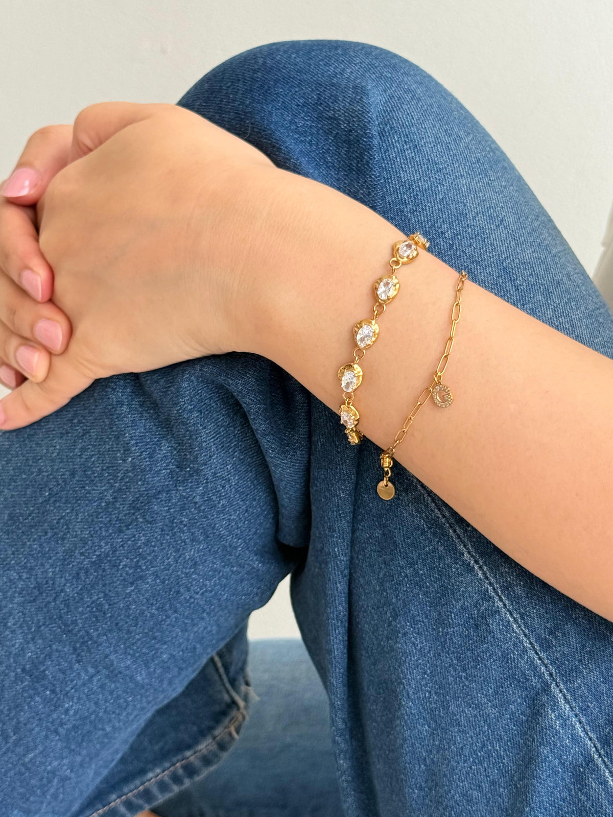 A close-up of a person's wrist wearing two delicate gold bracelets, one with zircon stones and one with small gold-plated initial pendant, styled with blue denim jeans.