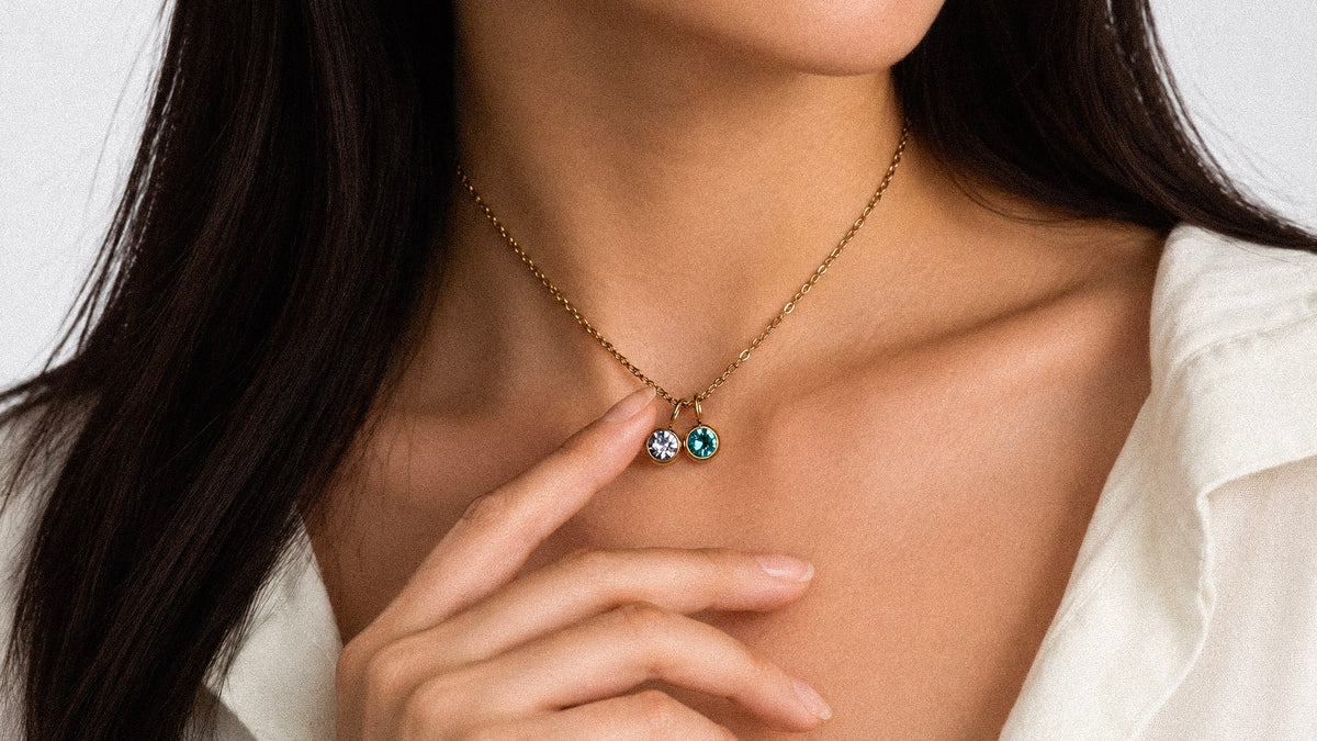 A close-up of a woman wearing a delicate gold chain necklace with two small round blue-green gemstone pendants, styled with a white collared shirt.