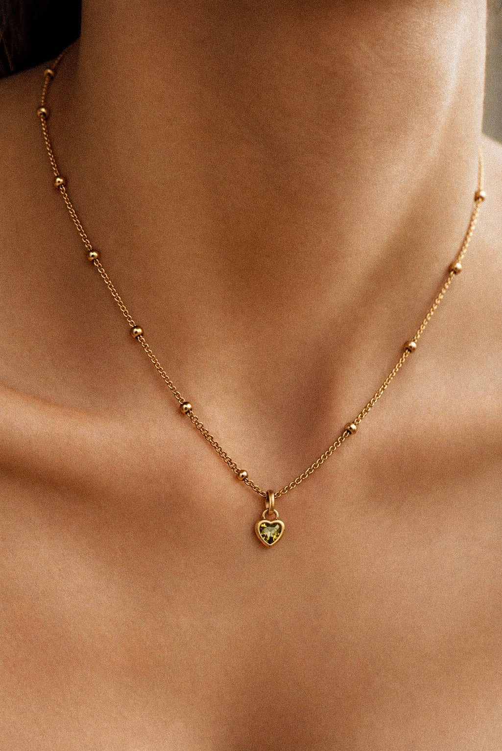 A delicate gold chain necklace with small ball accents and a small heart-shaped pendant featuring a green inlay, shown worn on a model's neck. august heart birthstone, 18K gold-plated, stainless steel necklace, non-tarnish, water-resistant