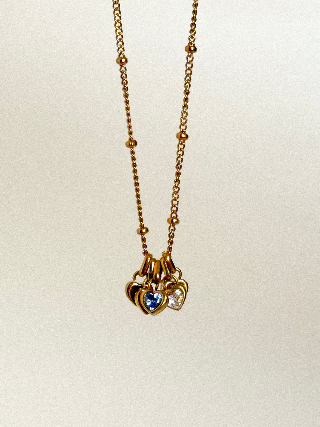 A gold chain necklace with a cluster of three heart-shaped charms, including a central blue heart and a right-side clear heart, shown against a plain light background.