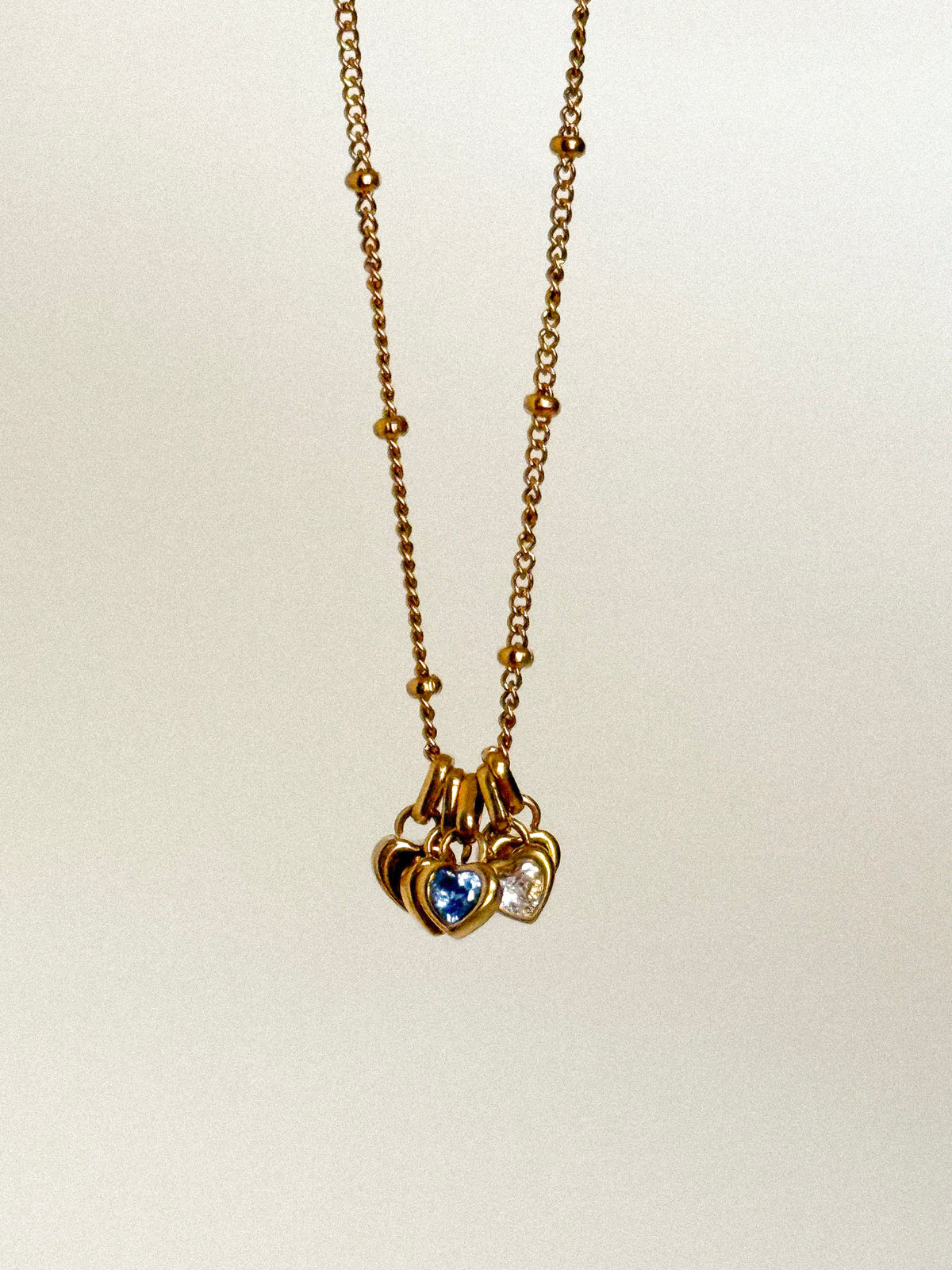 A gold chain necklace with a cluster of three heart-shaped charms, including a central blue heart and a right-side clear heart, shown against a plain light background.