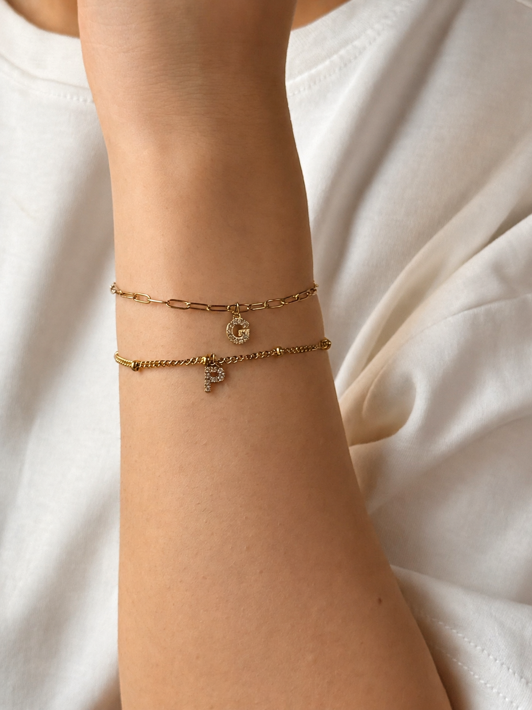 A close-up of a person's wrist wearing two delicate gold bracelets, one with a paperclip link design and one with a small charm, against a soft white top background.