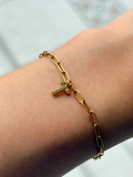 A gold paperclip chain bracelet with a small initial pendant paved with clear stones, shown worn on a person's wrist.