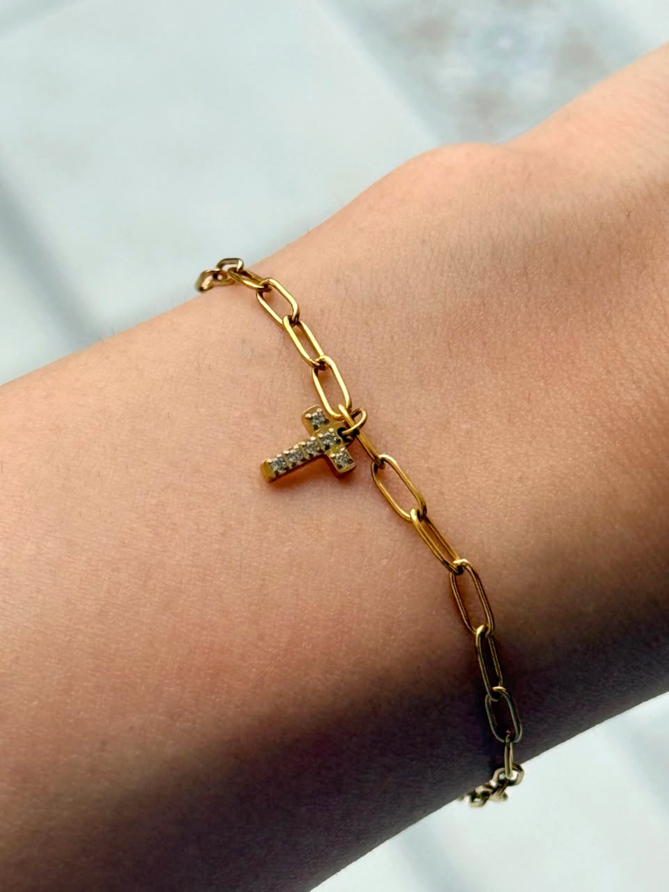A gold paperclip chain bracelet with a small initial pendant paved with clear stones, shown worn on a person's wrist.