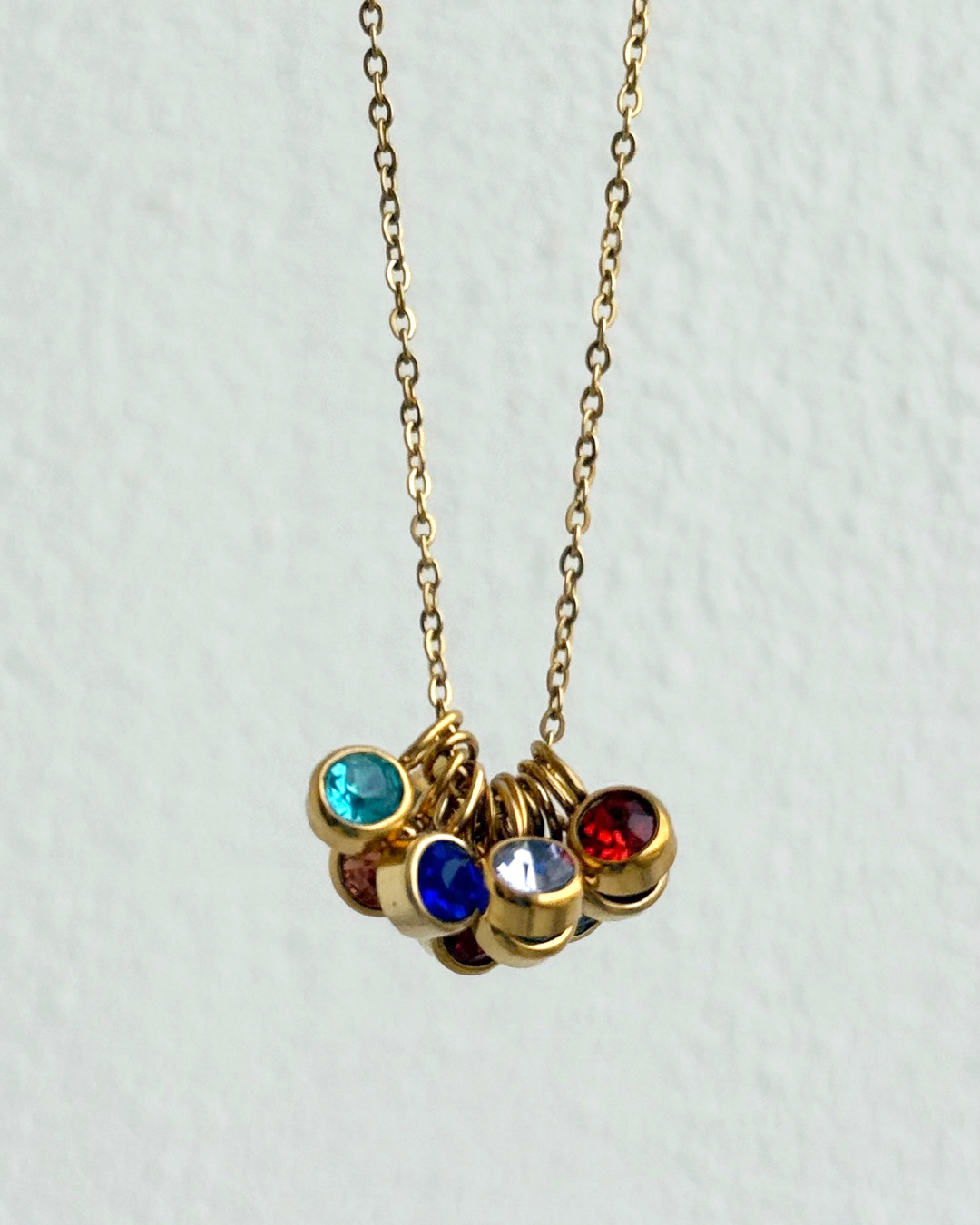 A gold chain necklace with a cluster of small round gemstone charms in blue, red, white, and brown, shown against a light textured wall.