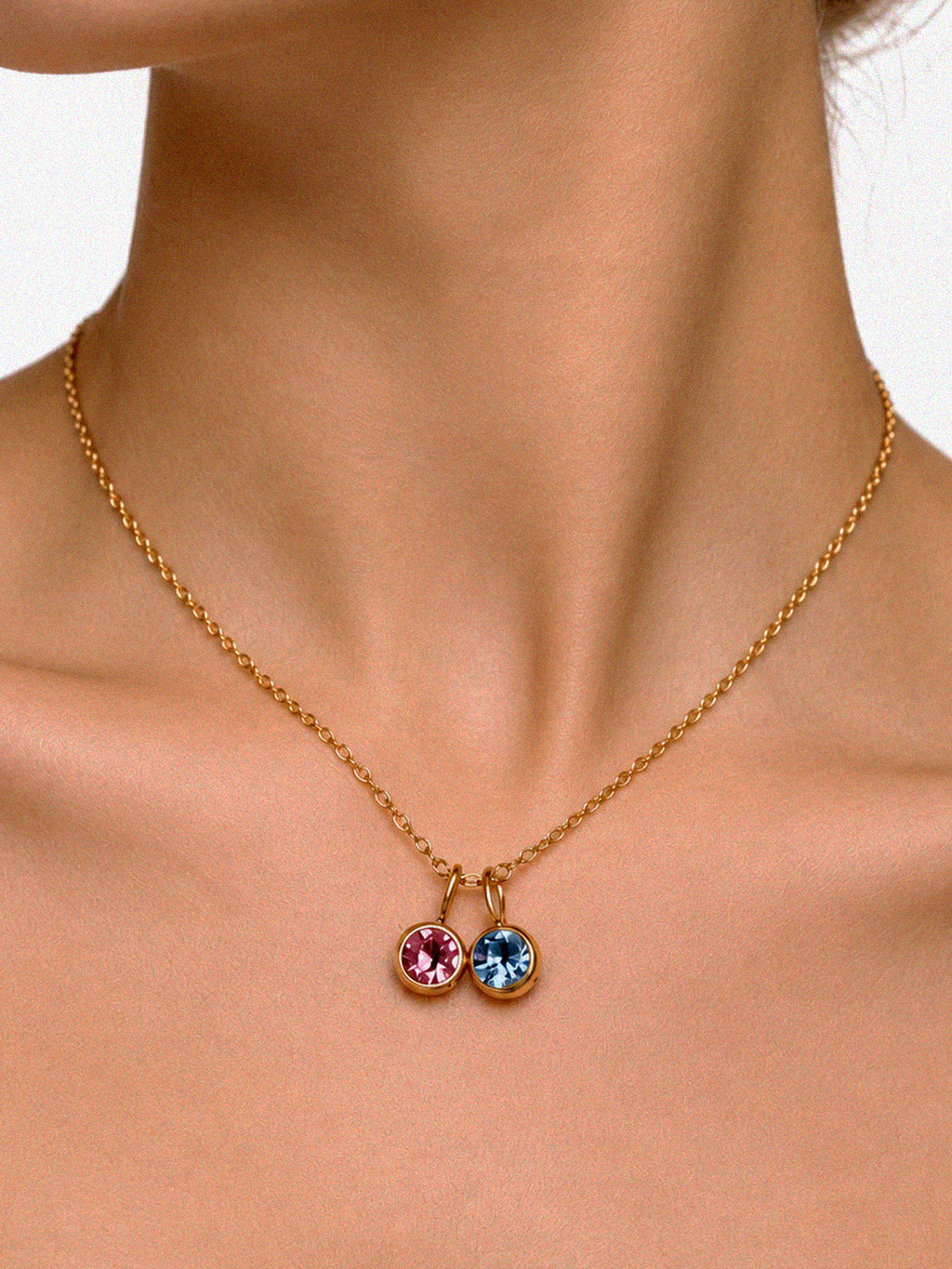 A close-up of a model's neck and collarbone wearing a delicate gold chain necklace with two small round bezel-set gemstone pendants, one pink and one light blue.