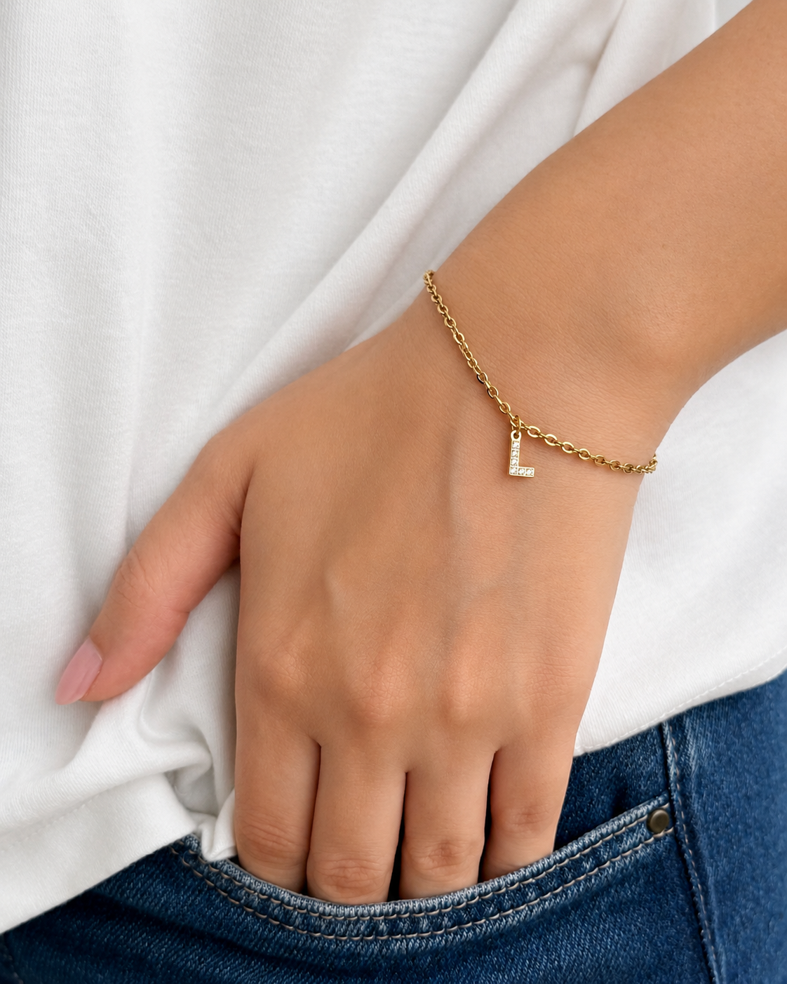 A close-up of a person's hand resting in a blue jeans pocket, wearing a delicate gold chain bracelet with a small initial 'L' charm.