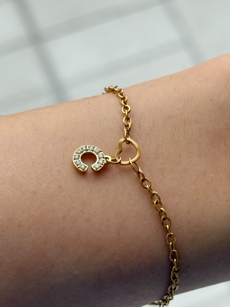 A gold chain bracelet with a small initial pendant charm set with clear stones, shown worn on a person's wrist.