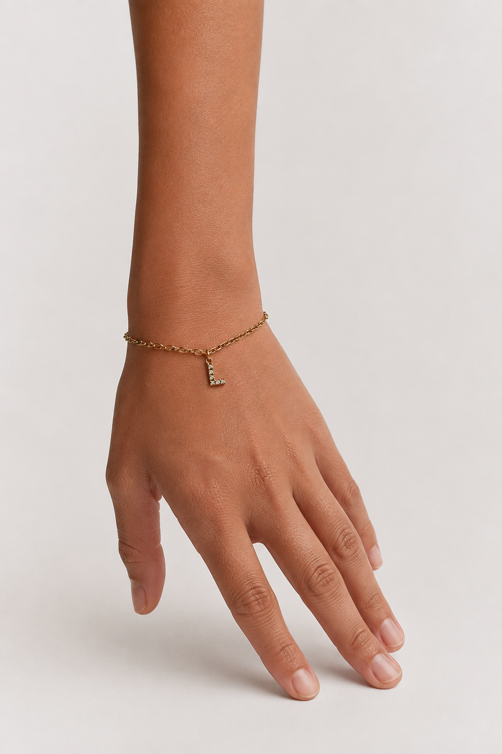 A delicate gold chain bracelet with a small letter 'L' charm embellished with clear stones, shown worn on a person's wrist against a light gray background.