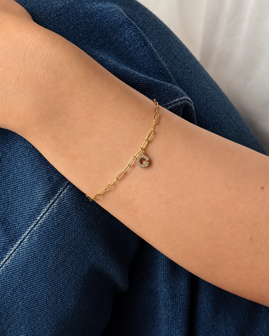 A delicate gold chain bracelet with a initial pendant featuring a clear stone, shown worn on a person's wrist over blue denim jeans.