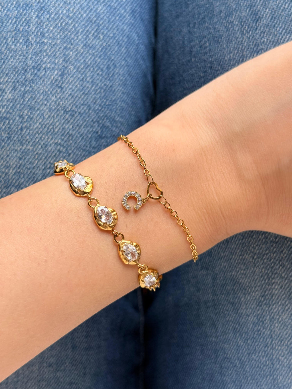 A gold bracelet with zircon stones and a small heart-chain with initial pendant, shown worn on a person's wrist against blue denim jeans.
